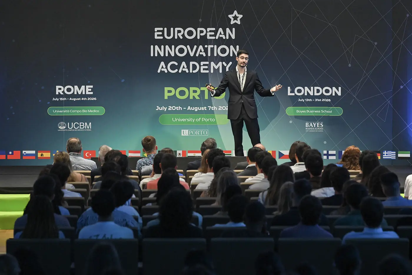 A speaker on stage in front of a large European Innovation Academy screen, addressing a full auditorium of students, with a row of national flags along the stage front.