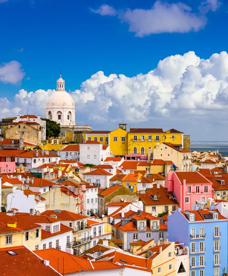 Terracotta rooftops of Lisbon's old town packed down a hillside beneath a white church dome, with the Tagus estuary beyond.