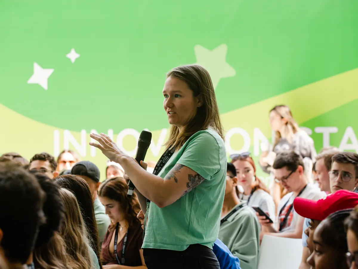 A participant with a microphone asks a question from a packed audience.