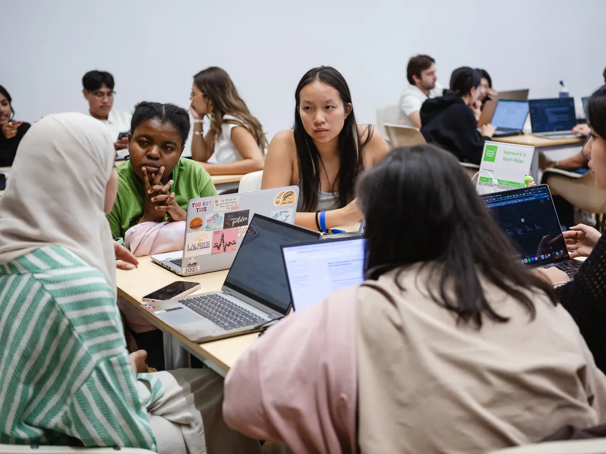 Students working in teams around tables with laptops, in a room filled with other student teams.